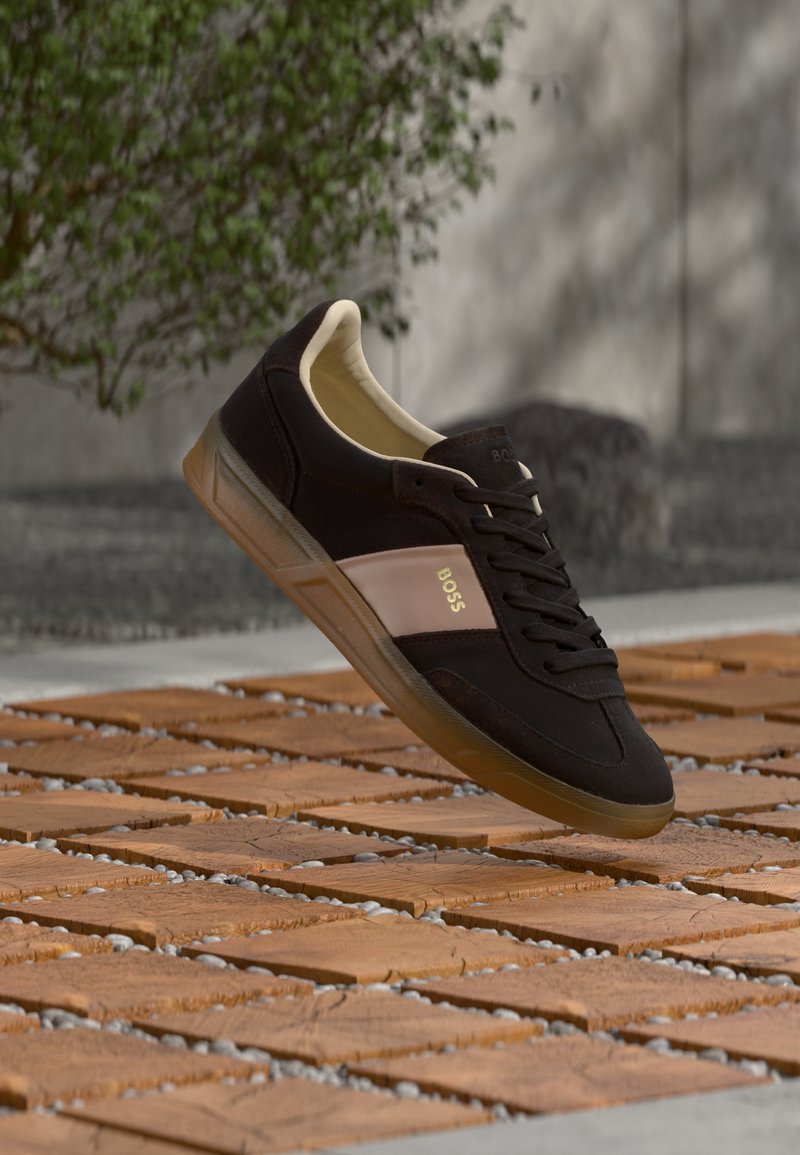 Baskets BOSS noires et beiges avec semelle en gomme flottant au-dessus du pavé en blocs de bois en plein air, à côté d'un buisson vert et d'un mur gris.