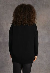 Person mit lockigem Haar, der ein schwarzes Langarmoberteil und schwarze Hosen trägt, steht gegenüber einer grau strukturierten Wand.