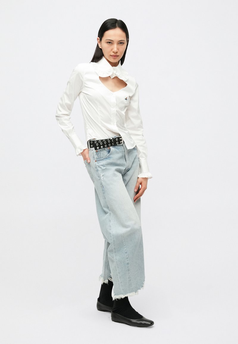 Vit skjorta med knappar, utskuren detalj och ryschkrage, kombinerad med ljusblå vida jeans. Svart skärp med cirkulära detaljer och svarta ballerinaskor.