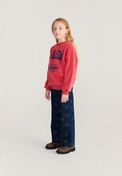 Enfant aux longs cheveux blonds debout, portant un sweat-shirt rouge avec un motif graphique, un pantalon bleu foncé avec des motifs floraux rouges, et des chaussures marron sur un fond uni.