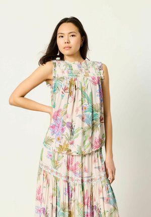 Kobieta ubrana w bezrękawnik w kwiatowy wzór i pasującą długą spódnicę w pastelowych kolorach, stojąca z jedną ręką na biodrze na tle jednolitego tła.