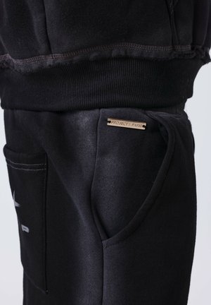Acercamiento al bolsillo de unos pantalones de chándal negros con una etiqueta metálica que dice "PROJECT X PARIS" y la cinturilla acanalada de la prenda negra correspondiente arriba.