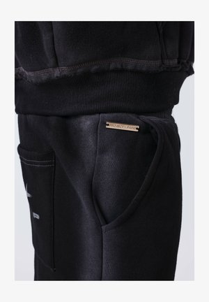 Close-up of black sweatpants pocket with metal tag reading "PROJECT X PARIS" and ribbed waistband of matching black garment above.