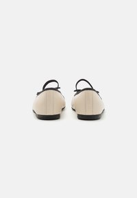Beige balletfeller med svarte detaljer, rund tå og en bakstropp. Glatt skinntekstur og subtil søm på sidene.