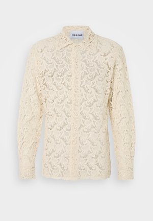 Krekls ar mežģīnēm krēmkrāsā, kas rotāts ar smalku ziedu rakstu, ar garām piedurknēm un klasisku apkakli ar pogu aizdari priekšpusē.