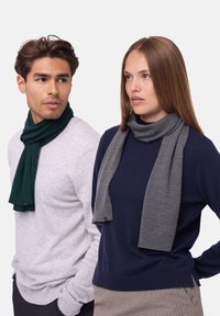 To personer, der bærer tørkl�æder: ét grønt og ét gråt. Tørklæderne er strikkede, bløde og hænger om deres hals. Casual stilarter og ensfarvede.