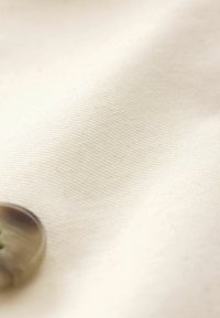 Katoenen stof in off-white met een fijn geribbeld textuur; een ronde, doorschijnende knoop is gedeeltelijk zichtbaar aan de rand.
