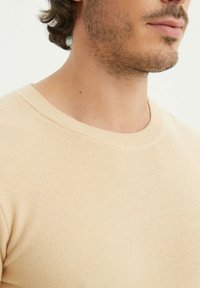 Pull en tricot beige clair avec un col rond, tissu texturé et détails en côtes subtils au niveau du col. Vue rapprochée.