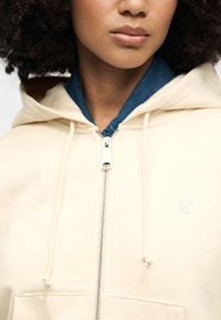 Μπεζ hoodie με φερμουάρ και κορδόνια, με μπλε γιακά από κάτω. Ορατά είναι τα λογότυπα στο στήθος και η υψηλής ποιότητας μηχανισμός φερμουάρ.