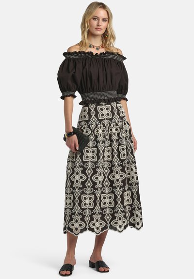 Femme portant un haut noir à épaules dénudées et une jupe midi noire avec broderie florale blanche, tenant une pochette noire et portant des sandales noires.