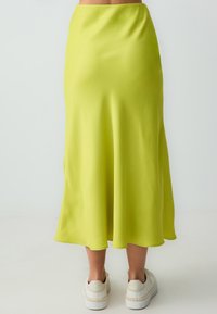 Limegrüner Satin-Midirock mit fließender, glatter Textur; verfügt über eine hohe Taille und einen schmal zulaufenden Saum, kombiniert mit weißen Sneakers.
