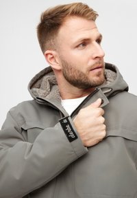 Grå, lynlåsjakke med hætte, der har et blødt, tekstureret foer og et sort label på manchetterne. Synlige stikninger som detaljer.