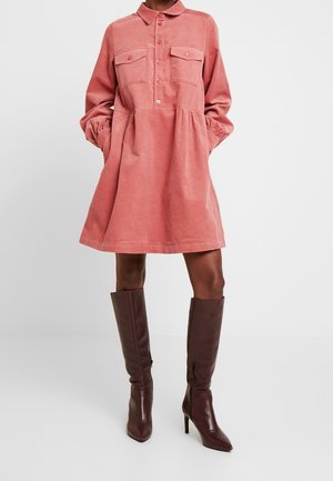 Robe en velours côtelé couleur rose doux avec un col chemise, fermeture à boutons, deux poches poitrine et manches longues. Associée à des bottes marron montant jusqu'aux genoux.
