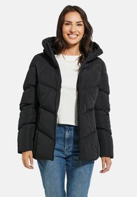 Svart puffjacka med huva, med en quiltad design, dragkedjestängning och sidofickor, som bärs över en vit topp och blå jeans.
