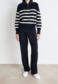Femme portant un pull à demi-fermeture éclair rayé bleu marine et blanc, un pantalon ample bleu marine et des baskets blanches, debout contre un mur blanc uni.