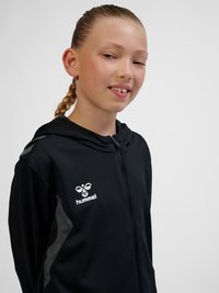 Junge Person mit geflochtenem Haar, die einen schwarzen Hummel Zip-Hoodie trägt, leicht lächelnd vor hellem, einfarbigem Hintergrund.
