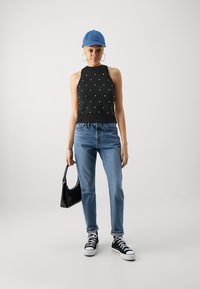 Svart halter-top med hvite prikker, blå denim jeans, svarte sneakers og en blå caps, holder en svart lakkhåndveske.