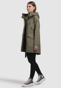 Veste d'extérieur vert olive avec capuche, fermeture éclair, deux poches avant et un tissu texturé. Portée sur des leggings noirs et des baskets.