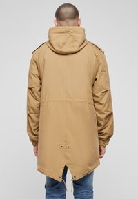 Giacca parka khaki con cappuccio, dotata di vita cinchata, maniche lunghe e orlo posteriore più lungo, realizzata in tessuto morbido e leggero.