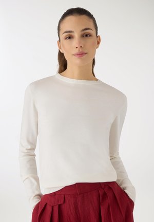 Giovane donna con capelli castani legati indietro, che indossa un maglione bianco a maniche lunghe e pantaloni rossi a vita alta, posando contro uno sfondo bianco uniforme.