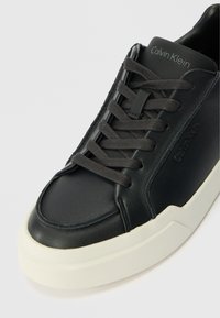 Fekete bőr sportcipő lekerekített orral, szürke fűzőkkel, textúrált talppal és oldalt látható, domború logóval.