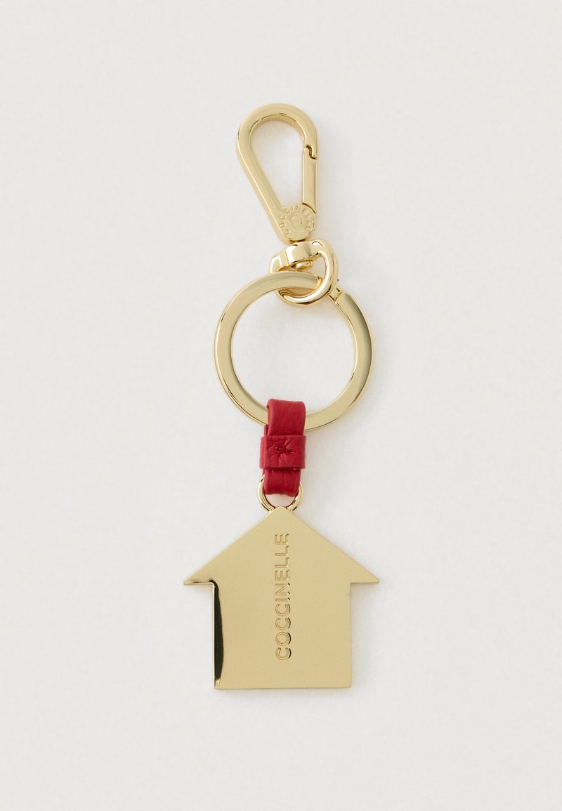 Porte-clés en forme de maison en or avec une sangle en cuir rouge et un fermoir rond, gravé de "COCCINELLE" sur un fond blanc.