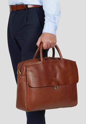 Mann i blå skjorte og mørke bukser holder en brun skinnmappe i håndtakene mot en ensfarget bakgrunn.