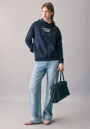 Sweat à capuche marine avec texte blanc, jean bleu clair, chaussures roses et sac à main noir. Le matériau semble doux ; design décontracté avec une poche frontale.