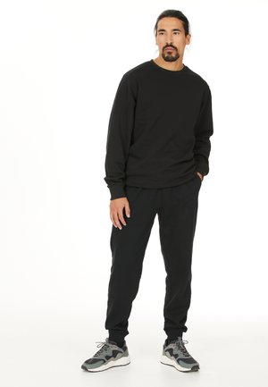 Zwarte lange mouwen sweatshirt met een ronde hals, gecombineerd met zwarte joggers. Het model draagt grijze en zwarte sneakers met een gestructureerde zool.