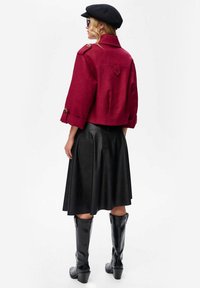 Rode cropped jasje met knoopdetails, zwarte leren rok en zwarte kniehoge laarzen. Model draagt een zwarte pet en zonnebril.
