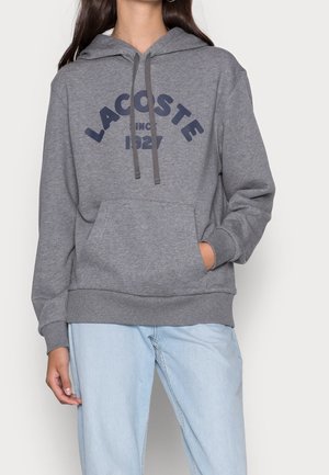 Persoon draagt een grijze Lacoste hoodie met de tekst "Lacoste Since 1927" en lichtblauwe jeans, met één hand in de zak.