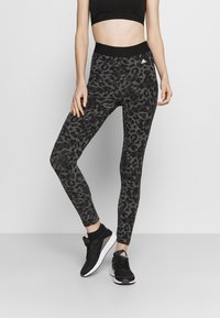 Leggings à motif léopard noir avec une taille haute, fabriquées en matériau extensible. Associés à des chaussures de sport noires avec des accents blancs.
