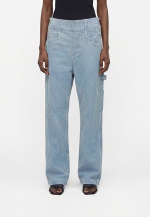 Osoba nosiaca nohavice s vysokým pásom, svetlomodré a bielo pruhované, s bočnými vreckami, širokými nohavicami a čiernymi topánkami s otvorenými prstami.