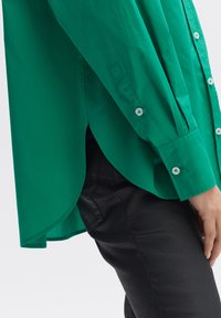 Groene button-up shirt met lange mouwen, met een gebogen zoom en contrasterende witte knopen, gecombineerd met zwarte strakke broeken.