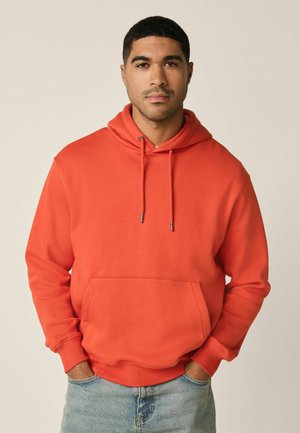 Man met kort zwart haar, gekleed in een oranje hoodie met een borstzak en lichtblauwe spijkerbroek, handen in de zakken, neutrale achtergrond.