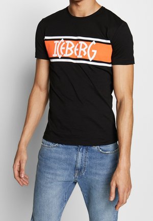 Homme portant un T-shirt noir à manches courtes avec un logo rayé orange, blanc et noir portant l'inscription "ICEBERG" sur la poitrine, associé à un jean bleu.
