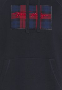 Felpa blu navy con un patch scozzese rosso e blu, con un logo rosso ricamato. Include una tasca frontale a kangaroo e cappuccio con coulisse.
