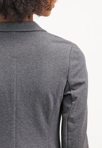 Blazer gris avec une texture de tissu lisse, doté d'un col cranté et d'une coupe ajustée. Les coutures visibles et les pinces aux épaules renforcent le design élégant.