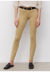 Béžové skinny džíny s hladkým povrchem, pěti kapsami a černým opaskem se stříbrnou sponou. Nosené s černými nazouvacími mokasíny.