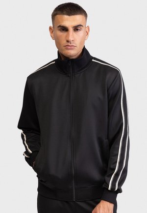 Čierna bunda na zips vyrobená z hladkej látky. Má biele pruhované detaily na rukávoch a bočných vreckách. Dizajn s vysokým golierom.