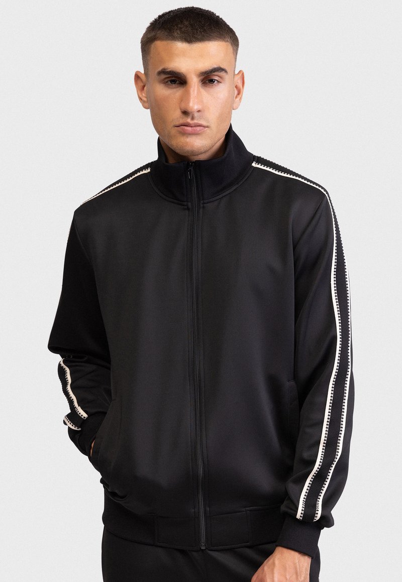 Μαύρο ζακέτα με φερμουάρ από λείο ύφασμα. Διακοσμημένο με λευκές ρίγες στα μανίκια και στις πλαϊνές τσέπες. Σχεδιασμός με ψηλό γιακά.