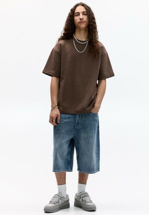 Bruine oversized katoenen t-shirt, denim shorts tot de knie, grijze sneakers. Accessoires omvatten gelaagde kettingen en ringen. Model staat rechtop.