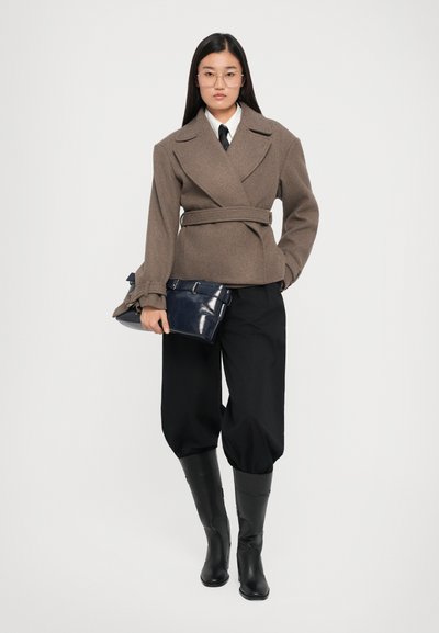 Brown, belted wool jacket with wide lapels, worn over a white shirt and black tie. Black, loose trousers and knee-high, smooth black boots.