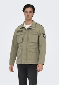 Giacca militare verde oliva con quattro tasche frontali, patch dell'esercito degli Stati Uniti e spalline. Tessuto di cotone con una vestibilità morbida.