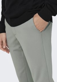 Pantalones ajustados gris claro con un frente plano, bolsillos laterales y una textura suave. Llevados con un suéter negro de canalé a la cintura.