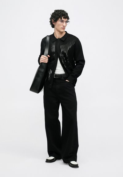 Cárdigan negro con detalles de lentejuelas, usado sobre una camisa blanca, combinado con pantalones anchos negros y zapatos negros con detalles blancos. Bolso negro de hombro.