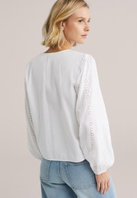 Blouse blanche à manches bouffantes, ornée de détails en broderie anglaise le long des manches. Associée à un jean bleu clair. Tissu en coton texturé.