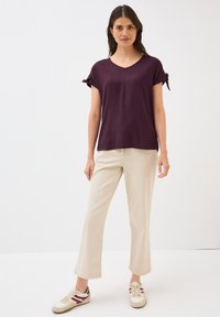 Haut violet à manches courtes avec un décolleté en V et des manches à nouer, associé à un pantalon beige clair et des baskets blanches avec des accents rouges. Texture de tissu lisse.