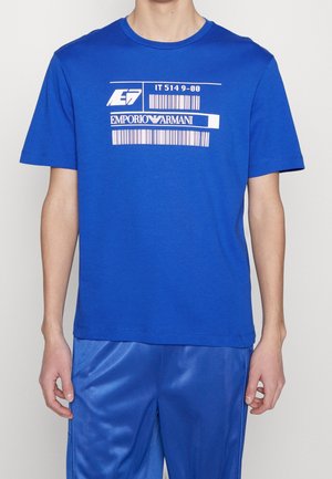 Mężczyzna w niebieskiej koszulce Emporio Armani z białym nadrukiem kodu kreskowego i napisem, w połączeniu z błyszczącymi niebieskimi spodniami.