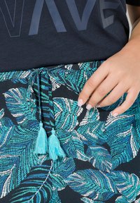 Jupe florale arborant des motifs de feuilles bleues et blanches sur un tissu noir, avec une cordelière ornée de deux pompons bleu clair.